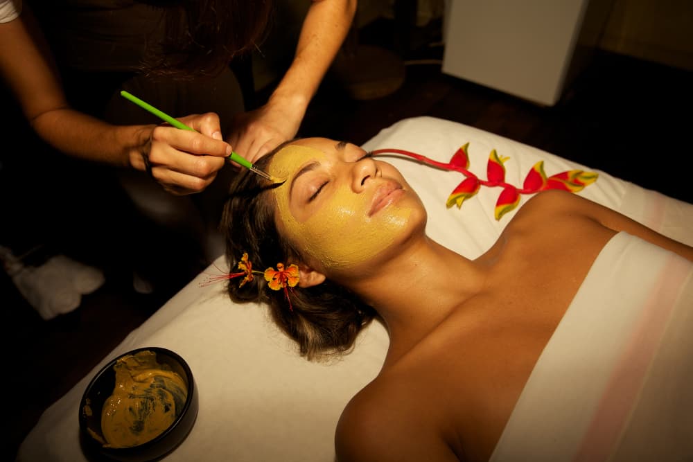 A woman lying on her back with her eyes closed, looking blissful, and getting her face brushed with a mask treatment
