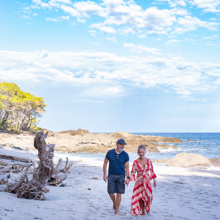 A couple walking hand in hand on the beach with lush greenery on one side and the beach on the other side.