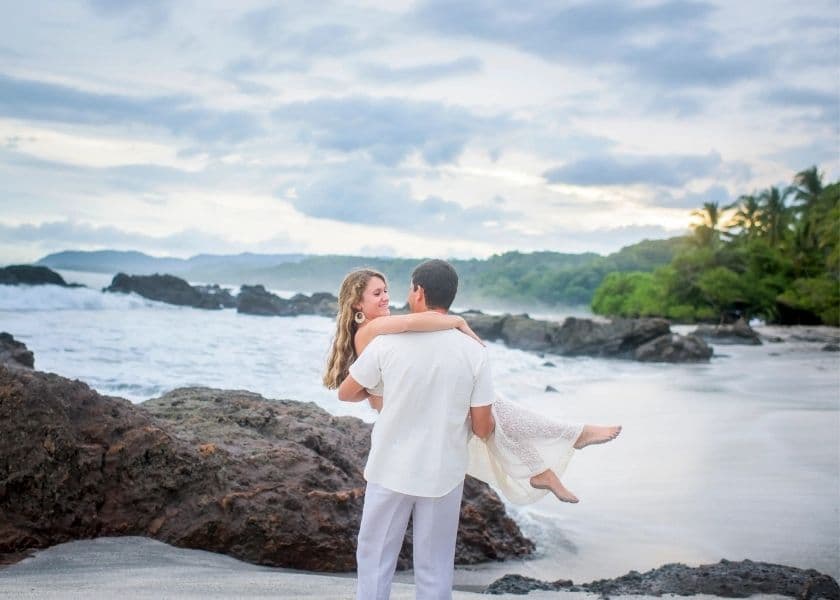 a groom carries his bride on a rocky beach with ocean waves and tropical greenery in the background at ylang ylang beach resort a groom carries his bride on a rocky beach with ocean waves and tropical greenery in the background at ylang ylang beach resort