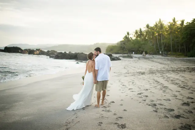 Couple in wedding attire walking hand in hand along a beach at sunset.