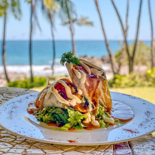 A plate featuring two stuffed tortillas drizzled with sauce, sitting on a bed of lettuce, against a backdrop of ocean and palm trees.
